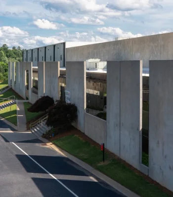 Modern concrete building exterior with tall rectangular columns, open side bays, grass, trees, a paved road, and a blue sky with clouds.