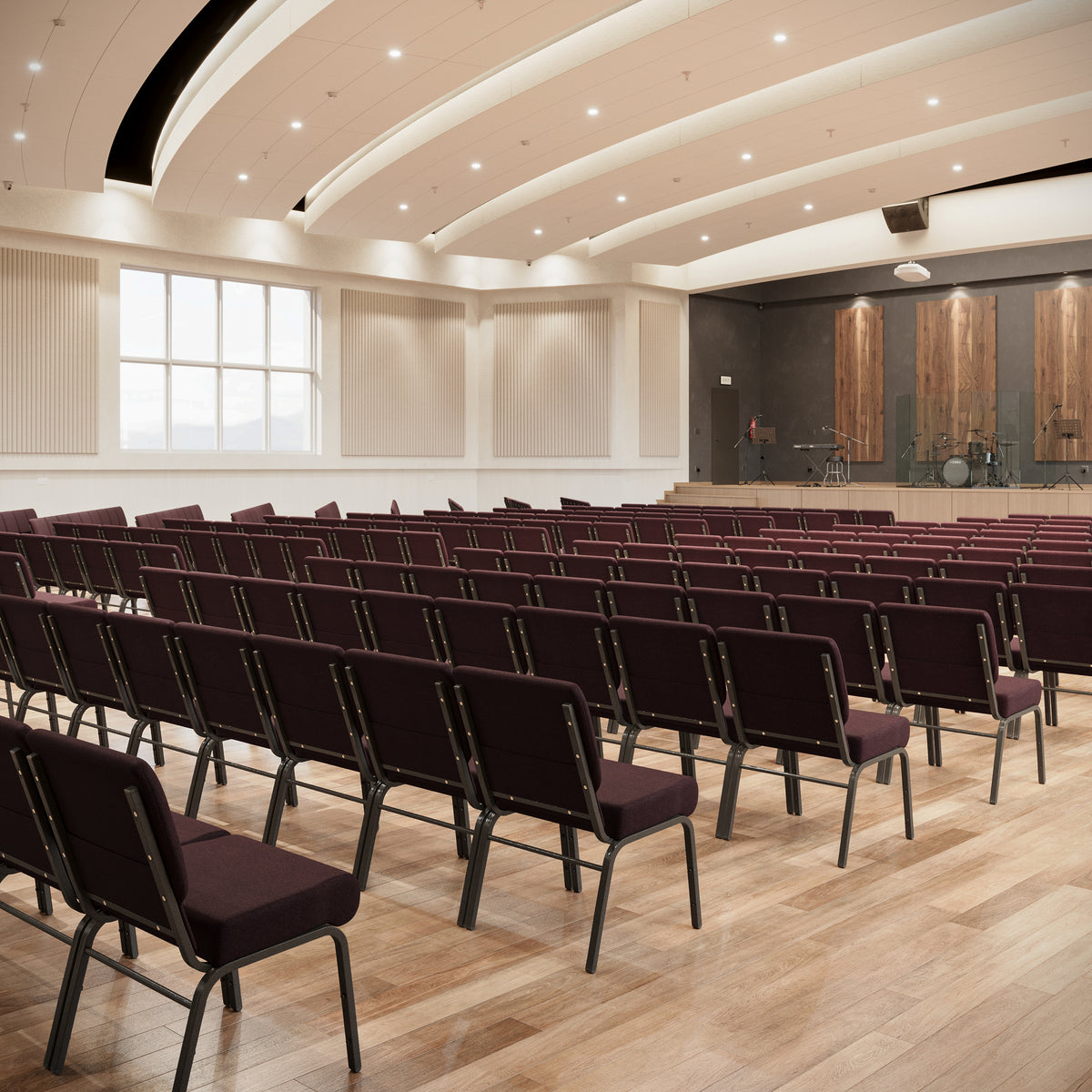 A large auditorium or lecture hall with rows of dark burgundy upholstered chairs facing a stage, set on a light wood floor with a curved cream ceiling and recessed lighting.